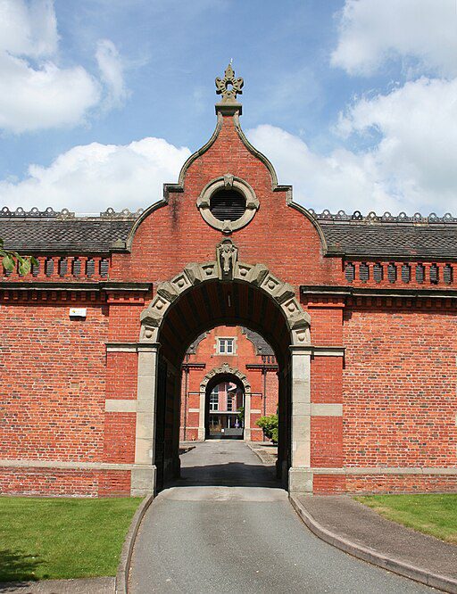 A beautiful red brick archway with decorative stonework stands proudly in Crewe, leading to a path lined with lush Cheshire greenery under a cloudy sky.